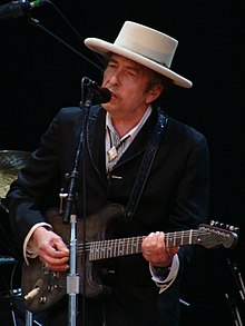 Bob Dylan plays a guitar and sings into a microphone.